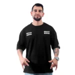 Un hombre con cabello corto y oscuro y barba lleva una camiseta negra personalizada de gran tamaño con el logotipo de Sons of Anarchy en texto blanco. Está de pie sobre un fondo blanco liso y hace gestos con las manos, capturando estilos de cultura pop.