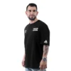 El hombre viste una camiseta negra con el logo de Sons of Anarchy - Camiseta Oversized y lleva un reloj en la muñeca, con la cara ligeramente girada hacia la izquierda sobre un fondo blanco impoluto.