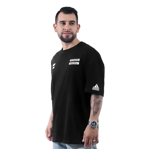 El hombre viste una camiseta negra con el logo de Sons of Anarchy - Camiseta Oversized y lleva un reloj en la muñeca, con la cara ligeramente girada hacia la izquierda sobre un fondo blanco impoluto.