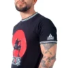 Una persona con barba, con una camiseta negra con un diseño gráfico que recuerda a estilos de anime y tatuajes en el brazo, se encuentra sobre un fondo blanco.