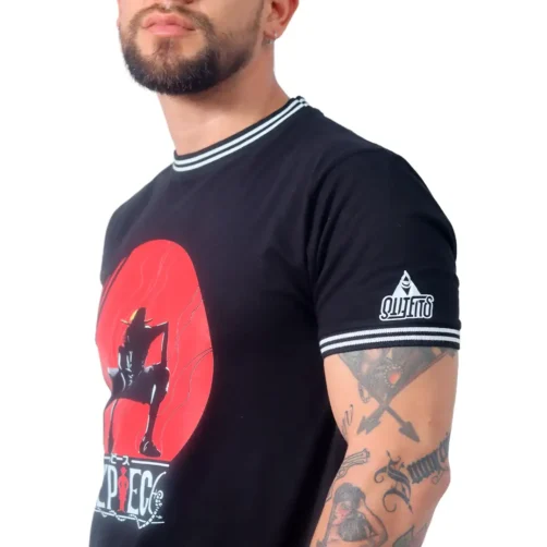Una persona con barba, con una camiseta negra con un diseño gráfico que recuerda a estilos de anime y tatuajes en el brazo, se encuentra sobre un fondo blanco.