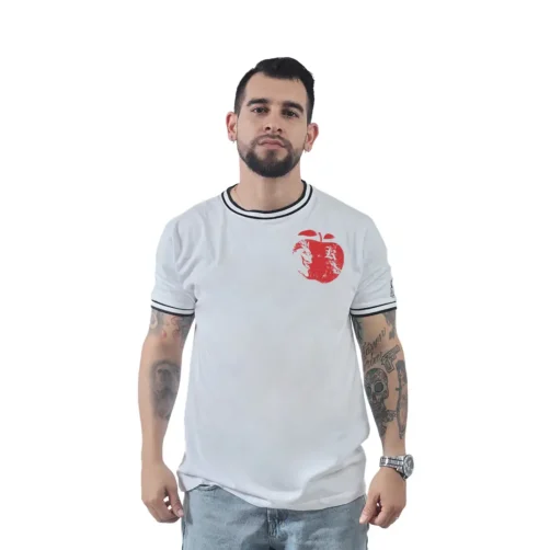 Un hombre con barba aparece de pie frente a un fondo sencillo, vistiendo una camiseta blanca con un diseño de manzana roja (un ejemplo perfecto de Camiseta Personalizada) combinada con jeans azules.