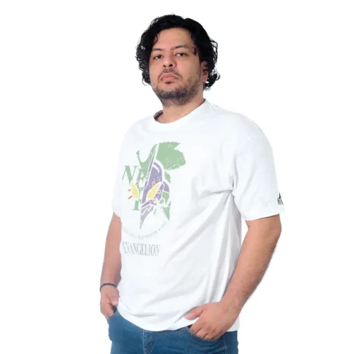Una persona con cabello rizado hasta los hombros y barba, que viste una camiseta blanca estampada con estilo retro y jeans, está parada sobre un fondo blanco.