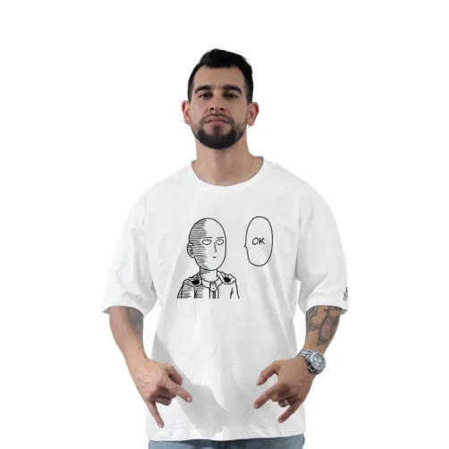 Una persona con barba que viste una camiseta personalizada, que muestra un personaje de dibujos animados y la palabra "OK" en estilos de anime, posa sobre un fondo blanco liso.