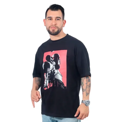 Un hombre con tatuajes en los brazos lleva una camiseta de gran tamaño de Star Wars, Cazador de recompensas, que muestra un gráfico de un personaje con casco sobre un fondo rojo que captura estilos de la cultura pop. Está de pie sobre un fondo blanco.