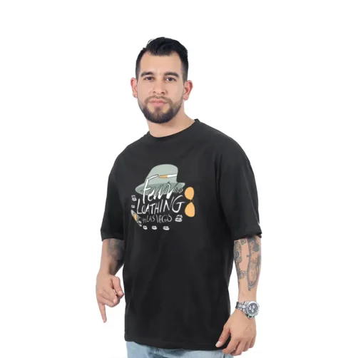Un hombre tatuado con barba viste la camiseta "Fear and Loathing in Las Vegas - Camiseta Oversized", con un diseño y texto retro, mientras señala con seguridad con su mano derecha.