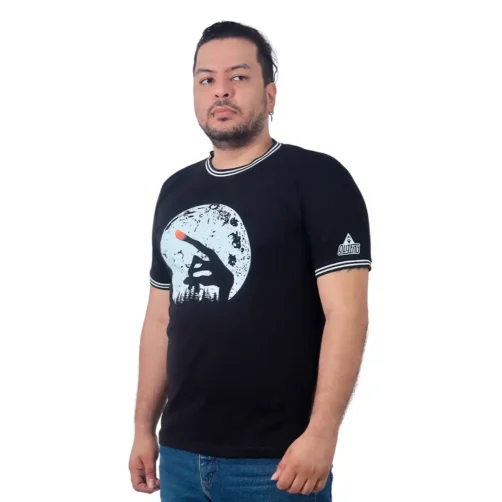 Un hombre que viste la camiseta "Dedo E.T. - Camiseta Cuello Tejido", que presenta un diseño de silueta iluminada por la luna, se encuentra sobre un fondo blanco liso, que representa estilos de cultura pop.