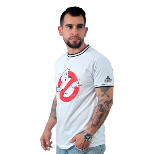 Un hombre con tatuajes viste una camiseta blanca de Los Cazafantasmas y unos vaqueros, encarnando a Estilos Gamer mientras mira hacia un lado contra un fondo liso.