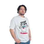 Una persona vestida con una camiseta "Stay Puft Cazafantasmas - Camiseta Cuello Tejido" y jeans azules se encuentra sobre un fondo blanco, capturando el estilo retro impregnado de estilo de cultura pop.