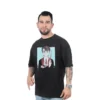 Un hombre con tatuajes lleva la camiseta de Harry Potter Anime - Camiseta Oversized, una camiseta negra que presenta un personaje de dibujos animados con gafas y una varita, que encarna estilos de cultura pop. Se para con confianza sobre un fondo blanco, lo que resalta su camiseta personalizada.