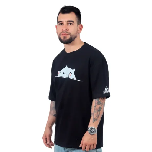Un hombre que viste una camiseta negra "Meme de Gatita - Camiseta Oversized" con un diseño de gato de dibujos animados se para con confianza sobre un fondo blanco.