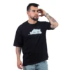 Un hombre con tatuajes, vistiendo una camiseta negra que dice "Meme de Gatita - Camiseta Oversized" y jeans, está parado con una mano en su cabeza, mirando hacia un lado.