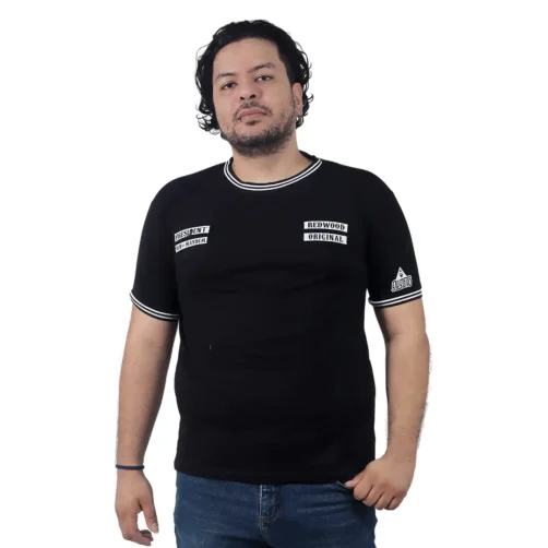 Una persona con cabello ondulado, vestida con un Logo de Sons of Anarchy - Camiseta Cuello Tejido y jeans azules, se encuentra sobre un fondo blanco liso, encarnando la esencia de la cultura pop.