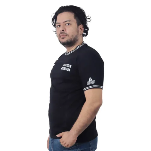 Un hombre de cabello oscuro y barba se encuentra de pie en el "Logotipo de Sons of Anarchy - Camiseta Cuello Tejido", que presenta un texto blanco en negrita que representa estilos de anime. Mira ligeramente hacia la cámara contra un fondo blanco liso, capturando una atmósfera relajada y relajada.